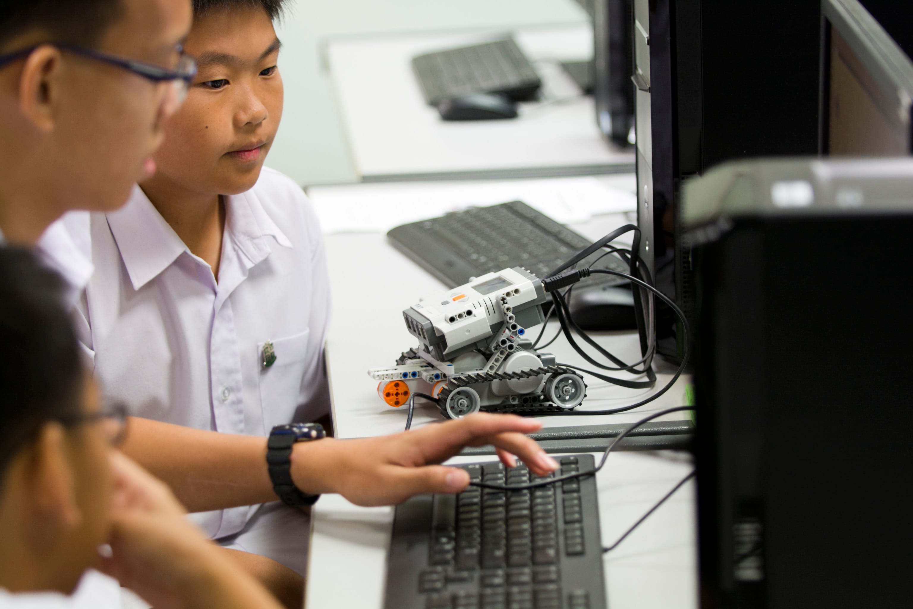 Two boys in white shirts work at a computer next to a small LEGO Mindstorms robot on a desk.